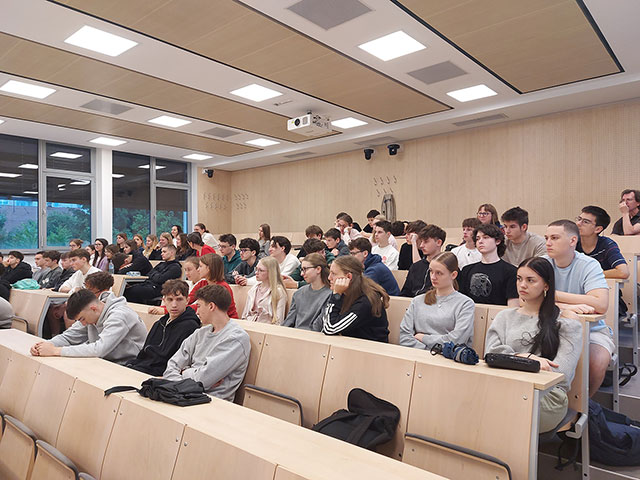 Pogled na polno predavalnico z lesenimi klopmi, kjer dijaki pozorno poslušajo predavanje.