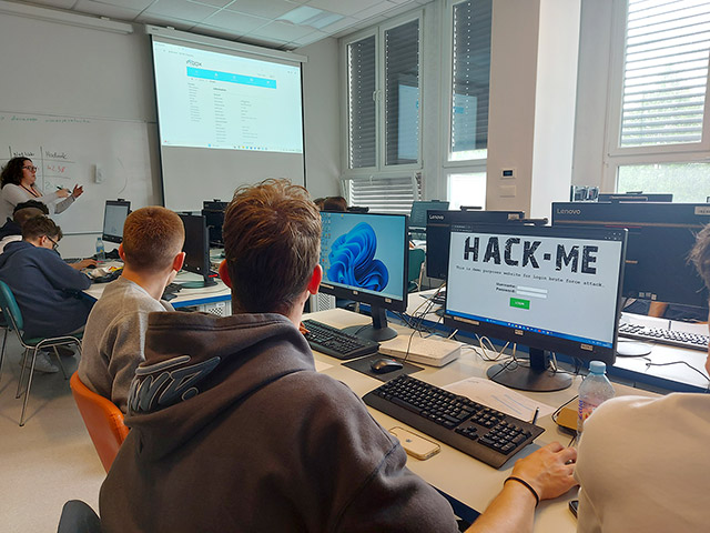 Pogled izza hrbtov dijakov v računalniški učilnici, kjer eden izmed monitorjev prikazuje spletno stran z napisom "HACK-ME", v ozadju pa predavateljica piše po tabli.