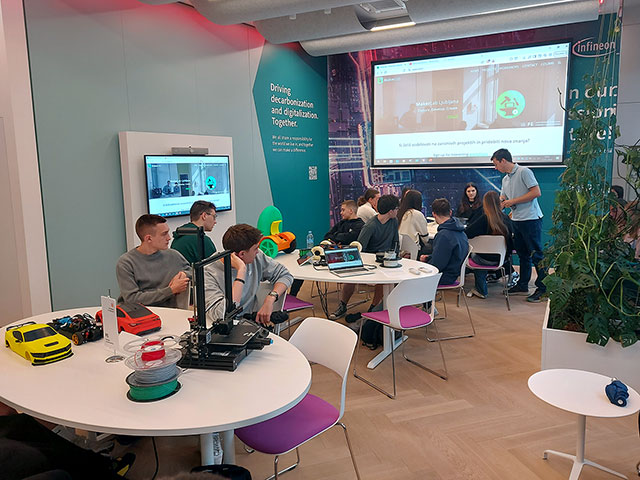 Dijaki sedijo okoli okroglih miz v sodobno opremljenem prostoru MakerLab, kjer si ogledujejo makete vozil, kolute z navito plastiko za tiskanje in 3D-tiskalnik.