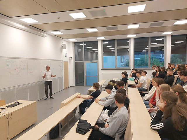 Profesor stoji pred šolsko tablo v veliki, svetli predavalnici in razlaga snov skupini dijakov, ki sedijo v klopeh.