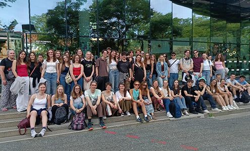 Skupinska fotografija večje skupine dijakov, ki pozirajo pred sodobno stavbo z veliko stekleno fasado, v kateri odsevajo okoliška drevesa. Nekateri sedijo na stopnicah, drugi stojijo za njimi.