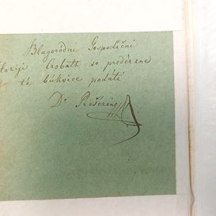 Bližnji posnetek ročno napisanega posvetila s podpisom »Dr. Prešerin« na svetlo zeleni podlagi, ki je vstavljena v staro knjigo z rjavimi platnicami.