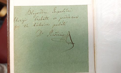 Bližnji posnetek ročno napisanega posvetila s podpisom »Dr. Prešerin« na svetlo zeleni podlagi, ki je vstavljena v staro knjigo z rjavimi platnicami.