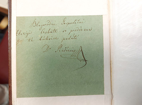 Bližnji posnetek ročno napisanega posvetila s podpisom »Dr. Prešerin« na svetlo zeleni podlagi, ki je vstavljena v staro knjigo z rjavimi platnicami.