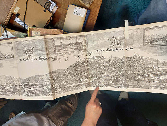 Pogled od zgoraj na veliko, raztegnjeno panoramsko grafiko Ljubljane (z napisom »Die Haupt Stadt Laybach«). V ozadju so vidne druge stare knjige.