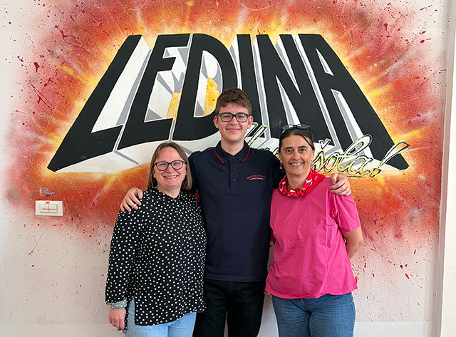 Dijak s prejšnje slike stoji v objemu med dvema profesoricama pred steno z barvitim grafitom "LEDINA".