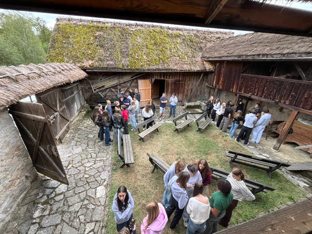 Pogled od zgoraj na kmečko dvorišče s tradicionalnimi lesenimi poslopji. Dijaki stojijo na dvorišču v treh večjih skupinah in se pogovarjajo.