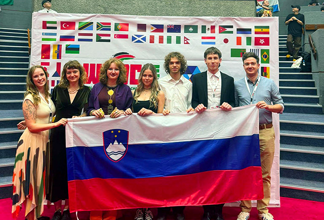 Člani slovenske debatne ekipe in mentor stojijo pred ozadjem z zastavami svetovnih držav in ponosno držijo slovensko zastavo.