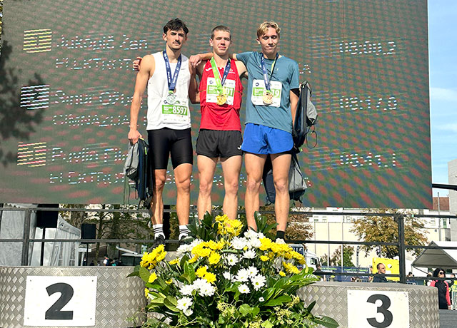 Na zmagovalnem odru stojijo trije tekači. Na majicah imajo startne številke, okrog vratu medalje. Oblečeni so v tekaška oblačila.