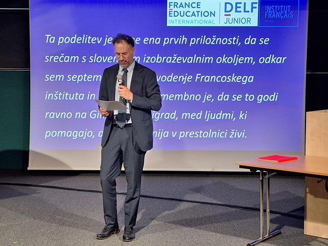 Moški v temno modri obleki s kravato stoji na odru pred velikim modrim zaslonom, drži mikrofon ter papir in govori na podelitvi diplom DELF.