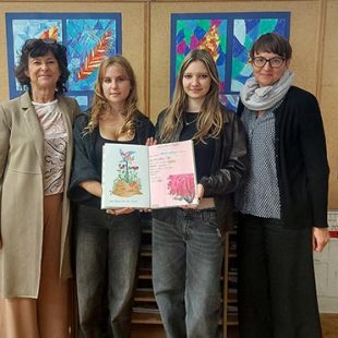 Dve učiteljici in dve dijakinji stojijo pred steno z otroškimi ilustracijami. Dijakinji v rokah držita odprt zvezek, v katerega so zapisana sporočila dijakov o miru.