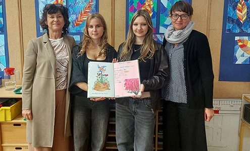 Dve učiteljici in dve dijakinji stojijo pred steno z otroškimi ilustracijami. Dijakinji v rokah držita odprt zvezek, v katerega so zapisana sporočila dijakov o miru.