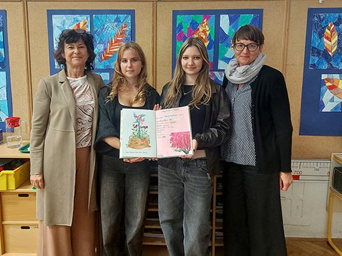Dve učiteljici in dve dijakinji stojijo pred steno z otroškimi ilustracijami. Dijakinji v rokah držita odprt zvezek, v katerega so zapisana sporočila dijakov o miru.